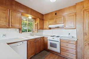 Kitchen featuring white appliances, light countertops, under cabinet range hood, dark wood-type flooring, and brown cabinetry
