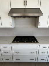 Kitchen with under cabinet range hood, stainless steel JennAir gas cooktop, white cabinetry, and light stone countertops