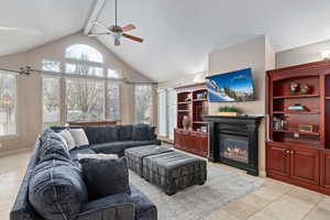 Living area featuring a glass covered fireplace, light tile patterned floors, beamed ceiling, high vaulted ceiling, and a ceiling fan