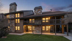 Back of house at dusk with a chimney, stone siding, a balcony, a lawn, and a patio area