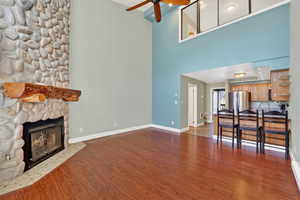 Living room with dark wood-style flooring, a ceiling fan, a stone fireplace, and a towering ceiling