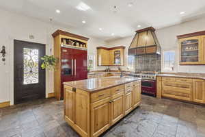 Kitchen with high end appliances, stone tile floors, custom range hood, an island with sink, and recessed lighting