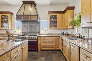 Kitchen featuring high end stove, premium range hood, stone finish floors, light stone counters, and glass insert cabinets