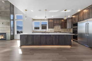 Kitchen with dark brown cabinetry, a kitchen island with sink, pendant lighting, built in appliances, and decorative backsplash