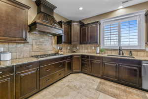 Kitchen with dark brown cabinetry, custom range hood, decorative backsplash, light stone countertops, and stainless steel appliances