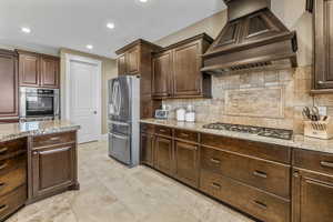 Kitchen featuring custom range hood, dark brown cabinetry, appliances with stainless steel finishes, recessed lighting, and light stone countertops