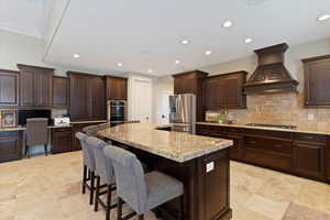 Kitchen with decorative backsplash, dark brown cabinets, custom exhaust hood, built in desk, and stainless steel appliances