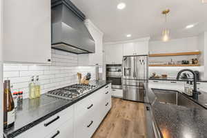 Kitchen featuring appliances with stainless steel finishes, custom range hood, dark stone counters, light wood finished floors, and recessed lighting