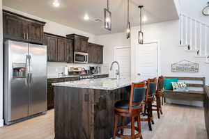 Kitchen with appliances with stainless steel finishes, dark brown cabinets, light stone countertops, a kitchen island with sink, and light wood-type flooring