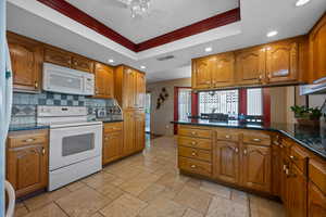 Kitchen featuring brown cabinetry, dark stone counters, white appliances, recessed lighting, and ornamental molding