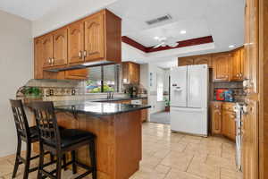 Kitchen with brown cabinetry, white appliances, decorative backsplash, a breakfast bar, and dark stone counters