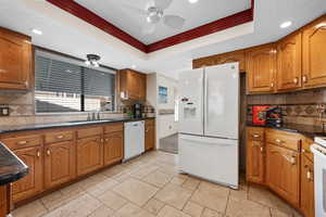 Kitchen featuring backsplash, ornamental molding, white appliances, brown cabinetry, and dark stone counters
