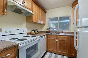 Kitchen featuring white appliances, under cabinet range hood, brown cabinetry, dark countertops, and a textured ceiling