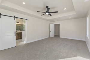Unfurnished bedroom with a tray ceiling, light colored carpet, recessed lighting, ceiling fan, and a barn door