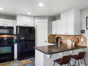 Kitchen featuring black appliances, dark countertops, backsplash, white cabinetry, and a peninsula