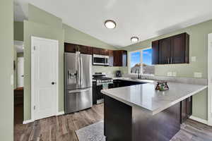 Kitchen with dark brown cabinetry, stainless steel appliances, a peninsula, lofted ceiling, and wood finished floors