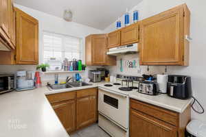 Kitchen featuring white electric stove, light countertops, brown cabinets, under cabinet range hood, and stainless steel microwave
