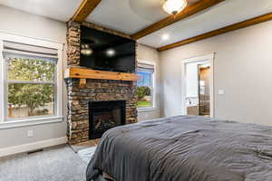 Carpeted bedroom featuring beamed ceiling, a stone fireplace, and ceiling fan