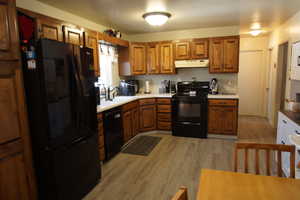 Kitchen featuring black appliances, brown cabinetry, light wood-type flooring, under cabinet range hood, and light countertops