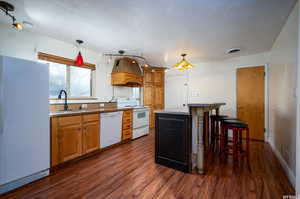 Kitchen featuring white appliances, a breakfast bar, dark wood-type flooring, brown cabinets, and custom range hood