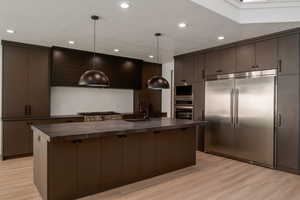 Kitchen with dark countertops, built in appliances, hanging light fixtures, a center island with sink, and recessed lighting