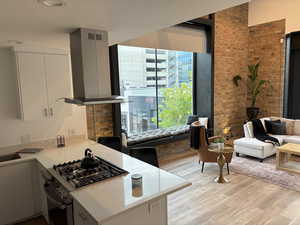Kitchen with stainless steel range with gas cooktop, wall chimney range hood, light wood-style floors, light stone countertops, and brick wall