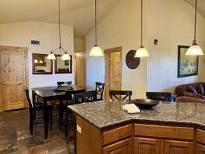Kitchen featuring brown cabinets, high vaulted ceiling, decorative light fixtures, dark stone counters, and a kitchen breakfast bar