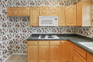 Kitchen featuring wallpapered walls, dark countertops, white appliances, light brown cabinets, and light tile patterned flooring