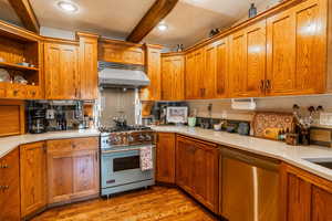 Kitchen featuring stainless steel appliances, light wood finished floors, range hood, beam ceiling, and brown cabinets