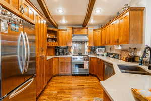 Kitchen featuring appliances with stainless steel finishes, brown cabinets, beam ceiling, open shelves, and light wood-type flooring