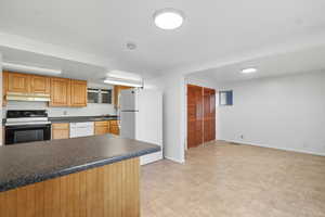 Kitchen featuring white appliances, dark countertops, under cabinet range hood, brown cabinetry, and a peninsula