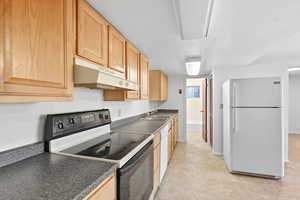 Kitchen with white appliances, light brown cabinets, under cabinet range hood, and dark countertops
