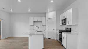Kitchen with stainless steel appliances, white cabinetry, a center island with sink, light stone counters, and light wood finished floors