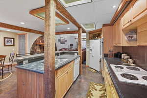 Kitchen featuring dark countertops, finished concrete floors, an island with sink, white appliances, and light brown cabinets