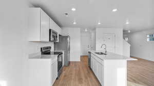 Kitchen featuring stainless steel appliances, white cabinets, a center island with sink, light wood finished floors, and light stone countertops