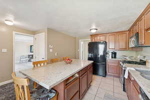 Kitchen with a breakfast bar, appliances with stainless steel finishes, a textured ceiling, light stone counters, and brown cabinets