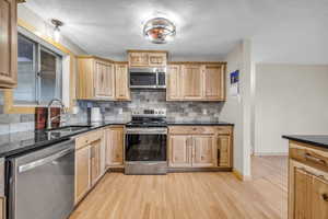 Kitchen featuring appliances with stainless steel finishes, backsplash, dark stone counters, a textured ceiling, and light wood-type flooring