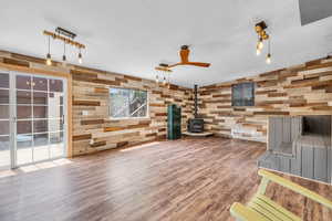 Unfurnished living room featuring a wood stove, wood finished floors, wooden walls, and ceiling fan