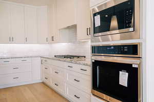 Kitchen with stainless steel appliances, light wood-style flooring, white cabinets, light stone countertops, and tasteful backsplash