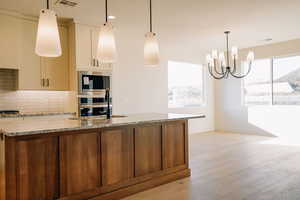 Kitchen with light stone countertops, backsplash, hanging light fixtures, an island with sink, and light wood finished floors