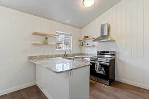 Kitchen with stainless steel range with electric stovetop, wall chimney range hood, a peninsula, light stone countertops, and open shelves