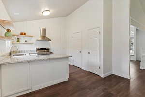 Kitchen with light stone counters, dark wood-type flooring, wall chimney exhaust hood, a peninsula, and high vaulted ceiling
