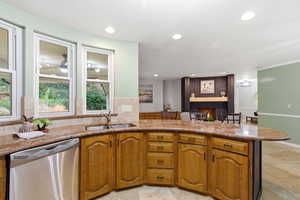 Kitchen with light stone counters, stainless steel dishwasher, a brick fireplace, brown cabinetry, and recessed lighting