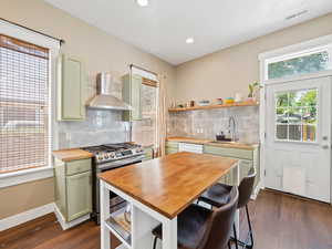 Kitchen with green cabinetry, backsplash, stainless steel gas range, butcher block counters, and wall chimney exhaust hood