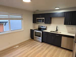 Kitchen featuring appliances with stainless steel finishes, light wood-type flooring, dark cabinetry, and light stone countertops