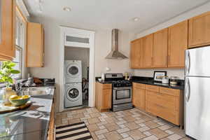 Kitchen featuring appliances with stainless steel finishes, wall chimney exhaust hood, stacked washer / dryer, recessed lighting, and dark stone countertops