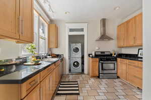 Kitchen featuring stainless steel appliances, wall chimney exhaust hood, dark countertops, stacked washing machine and dryer, and recessed lighting