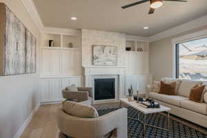 Living area with built in shelves, a stone fireplace, wood-type flooring, ornamental molding, and ceiling fan
