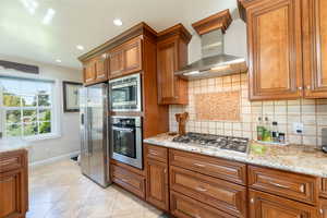 Kitchen featuring brown cabinets, backsplash, wall chimney range hood, stainless steel appliances, and light stone counters