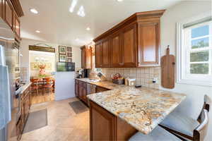 Kitchen with brown cabinets, backsplash, light stone counters, a breakfast bar area, and recessed lighting